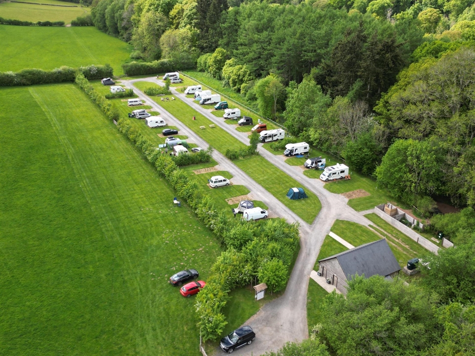 Drone photograph of Caravan site with shower block, fire pit, seating area and campers. Beautiful green landscape on private estate.