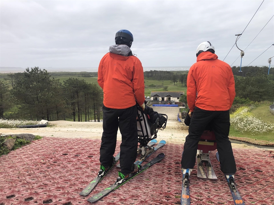 Two sit ski instructors in orange jackets each holding a sit ski are stood on top of an artificial ski slope preparing to ski.