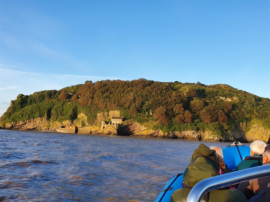 Steep Holm Island surrounded by water. People in boat looking at island which is steep and covered in trees and foliage. Remains of buildings can be s