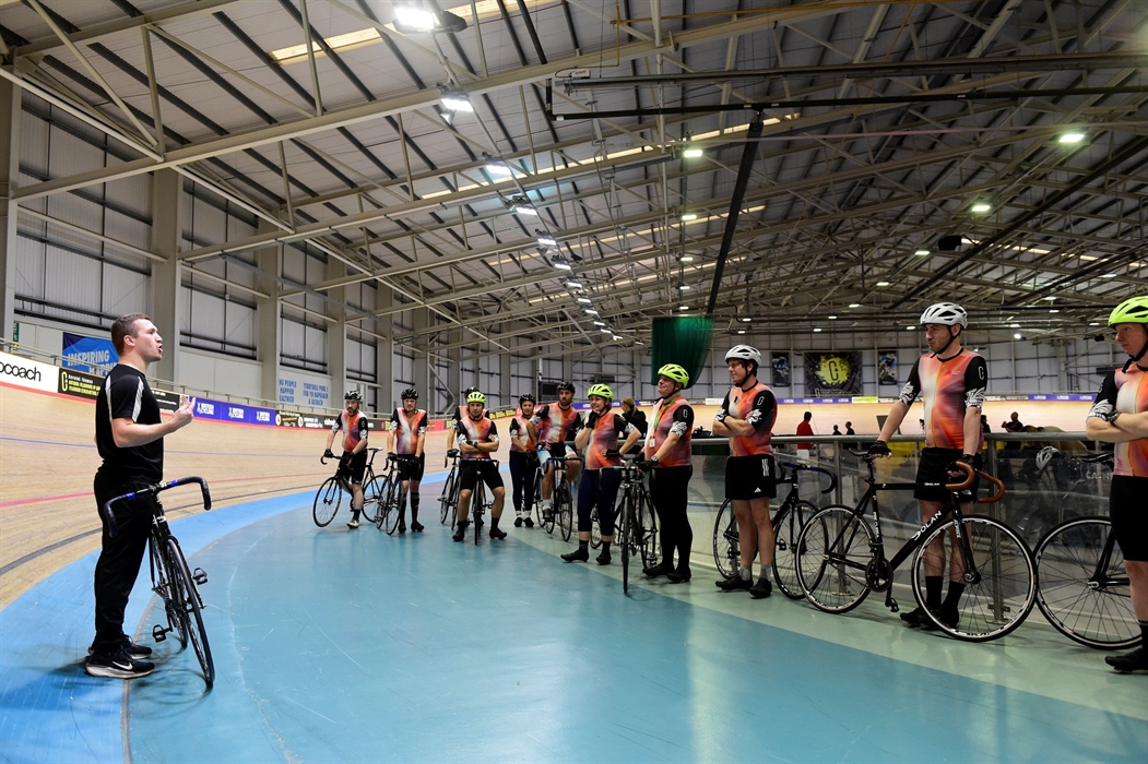 Group of people wearing cycling gear next to their bikes, listening to an instructor in a velodrome.