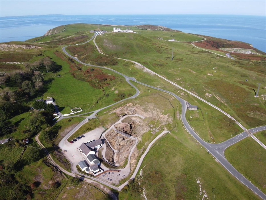 An aerial view, immediately below is the mine which looks like a beige landscape and white buildings that stand out from the green landscape beyond. B