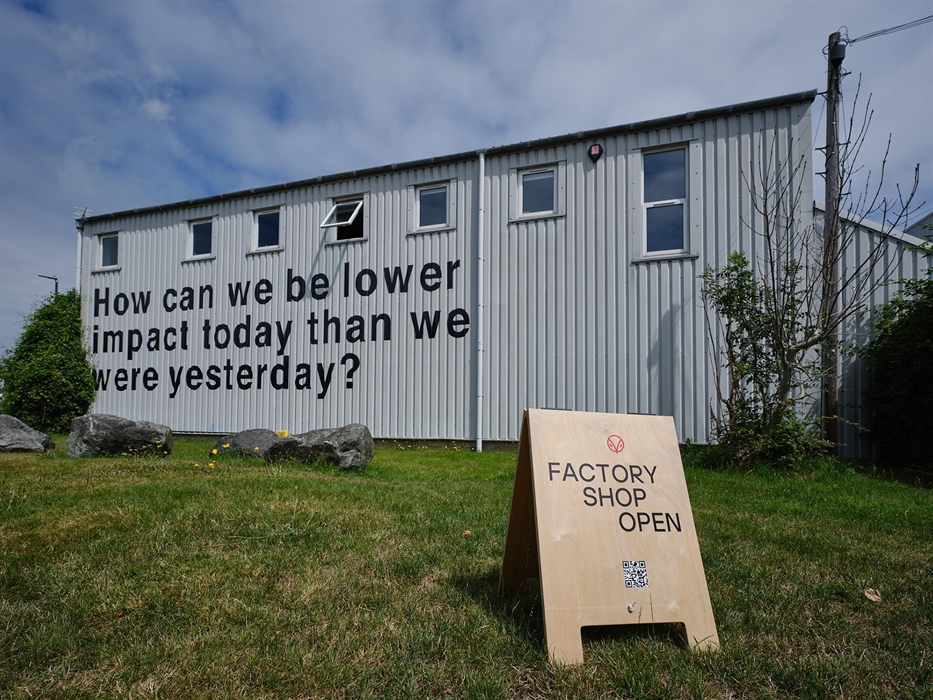 The exterior of the Hiut Denim factory features bold black text reading “How can we be lower impact today than we were yesterday?” A wooden sign on th