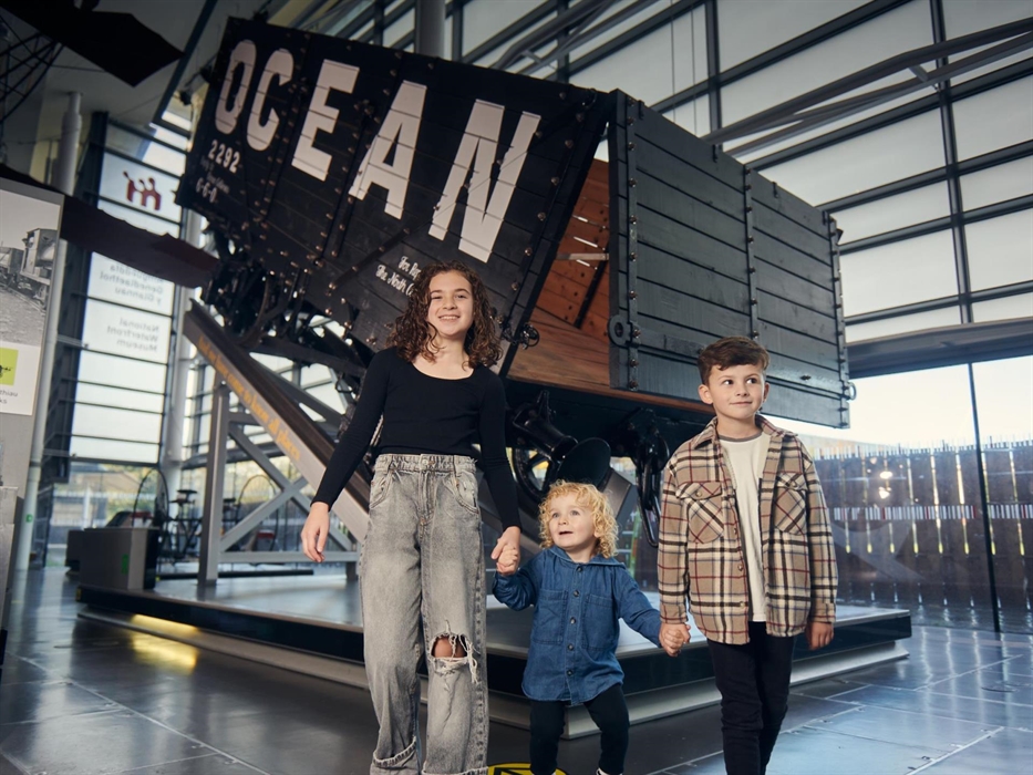a young girl and boy hold hands with a toddler as they explore a gallery full of old engines and industrial artefacts. Behind them is a large coal tru