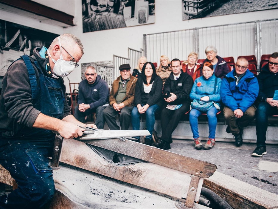 a craftsman in blue overalls sits on a bench working the piece of slate with a saw. in the background, a group of visitors are watching him. He explai