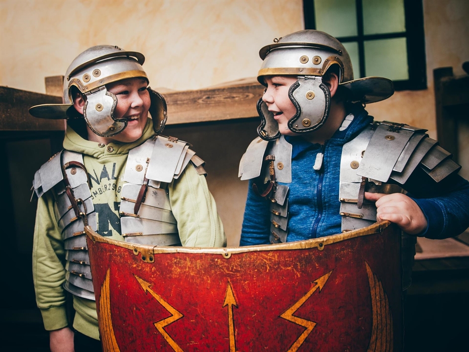Two young boys wearing Roman helmets and armour. They are holding up a red Roman shield and smiling.