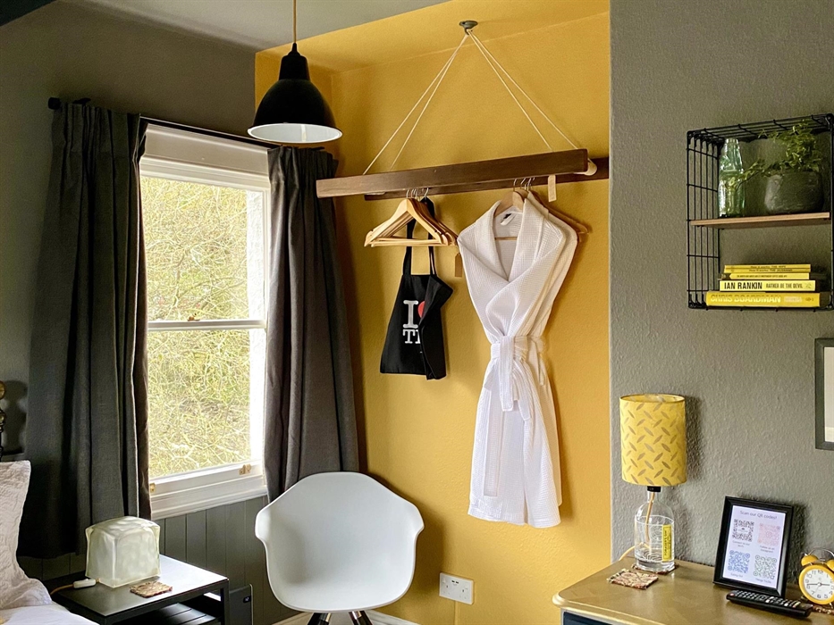 A yellow and grey bedroom, with a chair in the corner, chest of drawers and yellow lamp, and a bathrobe hanging on a yellow wall, at Crafnant House, B