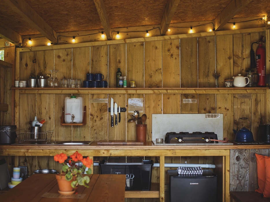 A camp kitchen with festoon lighting above, a countertop with shelving above with glasses and mugs on. A fridge is below the counter and in the foregr