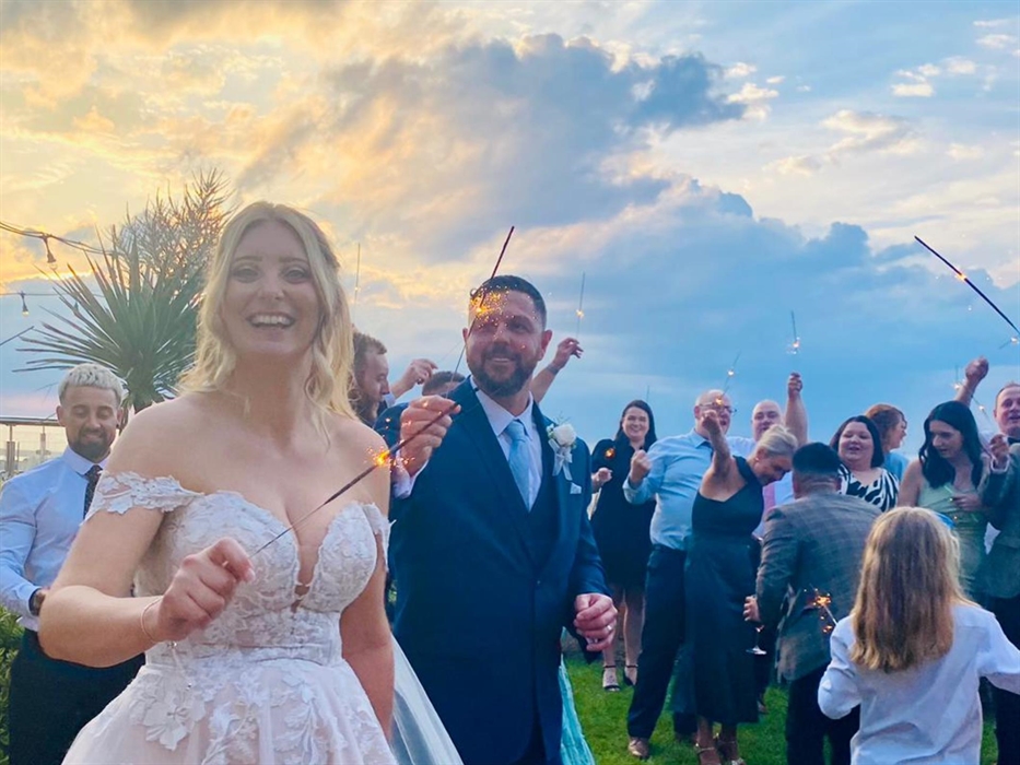 An image of a newly wedded bride and groom holding sparklers with their guests with the beach in the background, and the clouds golden from the sunset