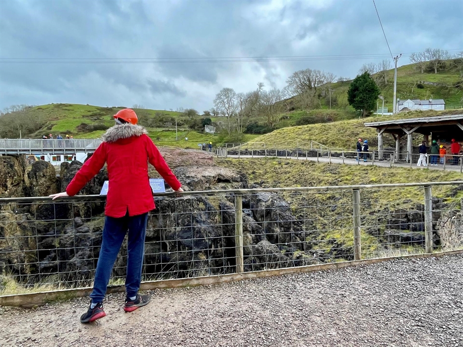 Person standing in foreground looking across a chasm in the ground towards a bridge where a small group of people stand.  On the right hand side of th