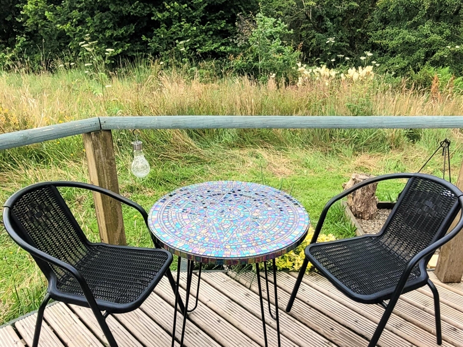 A handmade mosaic table in iridescent glass with 2 black woven chairs on the wood deck looking out to the meadow and the woodland behind.