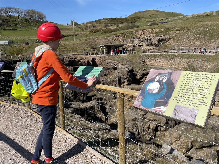 A child is standing reading an information board above a chasm in the rock below.  In the distance people are walking on the path on the other side an