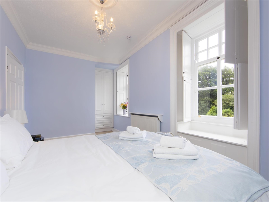 Light-filled double bedroom at Plas Cilybebyll with soft blue walls, a large sash window, and neatly folded towels atop crisp white bedding.