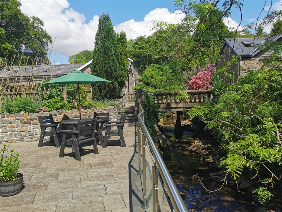 Black outdoor dining set on a paved terrace beside a stream, surrounded by stone walls and greenery