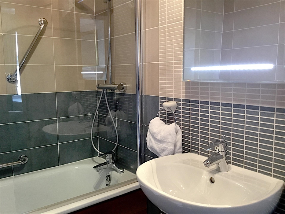 Double room en-suite bathroom showing a bathtub with a glass screen and shower, plus a wash basin with a lit mirror above it
