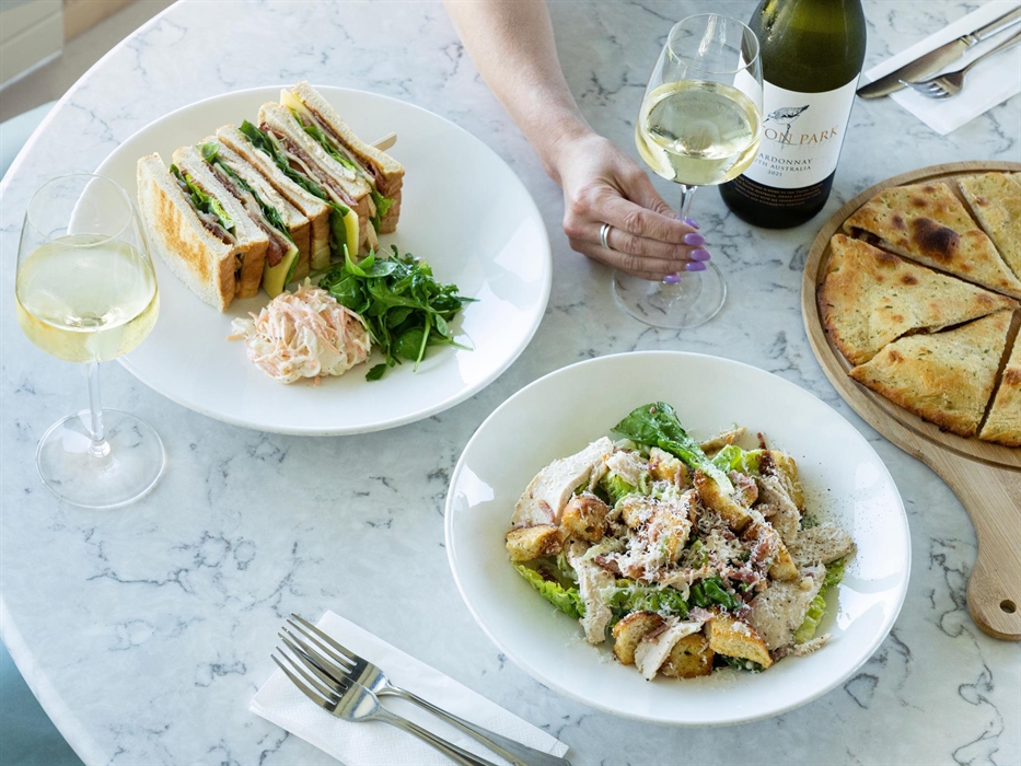Image of a woman's arm holding a glass of wine on a table with a plated club sandwich, another glass of wine, a chicken ceasar salad, and garlic bread