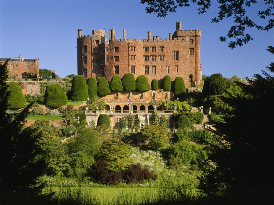 A red brick castle built on top of a hill with cascading terraces below and tall green yew tree hedges