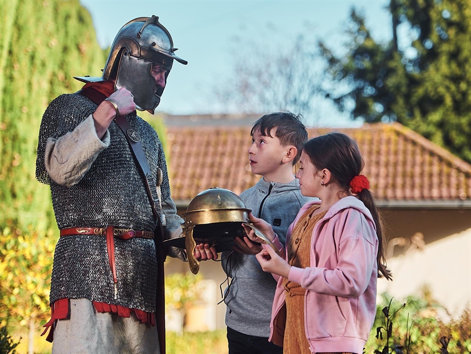 A young girl and boy are standing opposite a man dressed as a Roman gladiator. they are holding a Roman helmet and the gladiator is talking to the chi