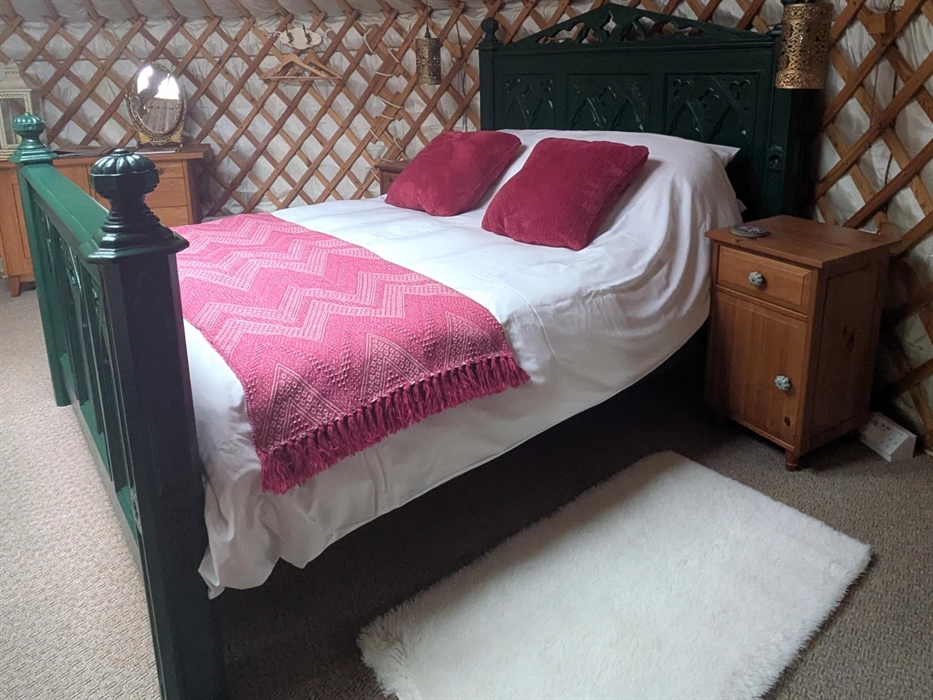 An ornate, tall, Gothic style, green, king-size bed . It has white bedding, a pink blanket and deep pink velvet cushions. There are wood bedside cabin