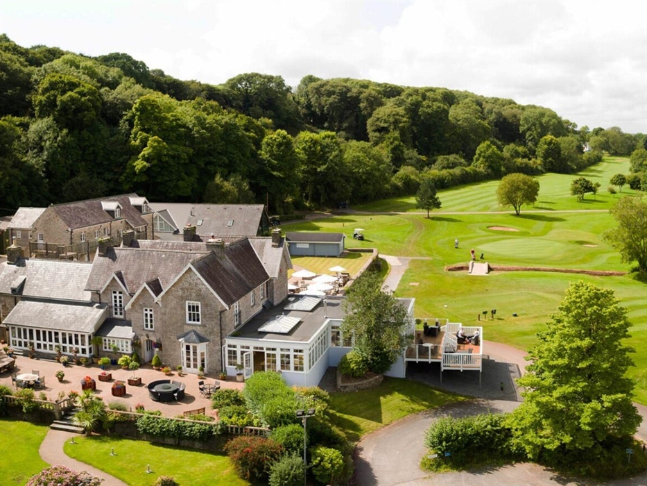 Charming stone manor nestled in lush countryside, with a sunlit terrace, conservatory, and golf course views. Surrounded by trees and rolling greens,