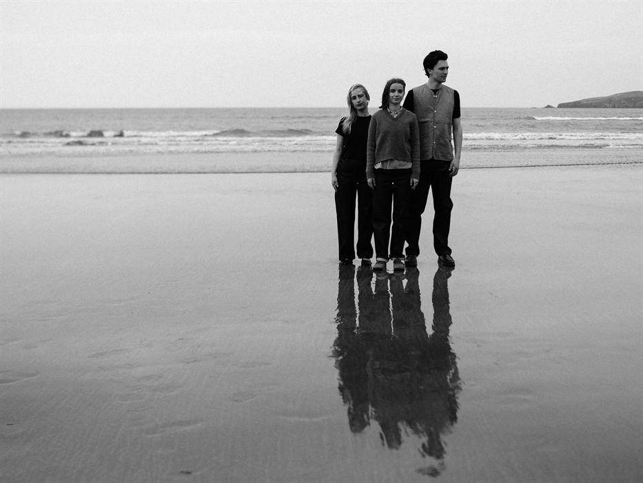 Three people stand on a wet beach facing slightly to the right, their reflections mirrored in the sand. The black-and-white photo captures a calm, coa