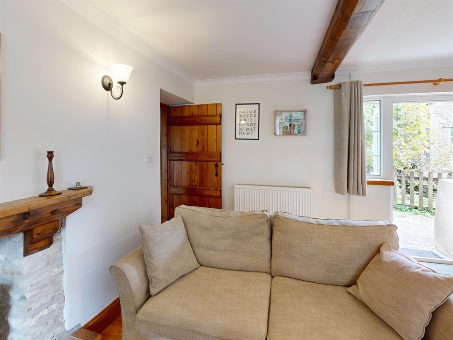 The inviting living room of Ty Cerbyd with light neutral furnishings, rustic wooden beams, and a door opening onto the garden.