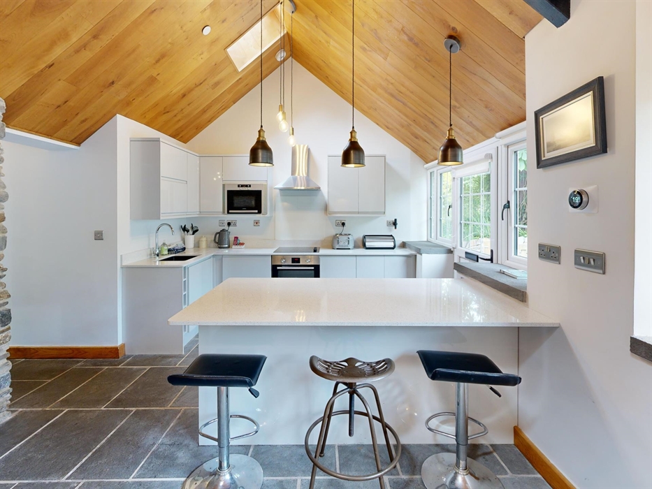 A spacious kitchen with white cabinets, slate flooring, a large island with stools, and pendant lighting beneath a vaulted wooden ceiling. A stone fea