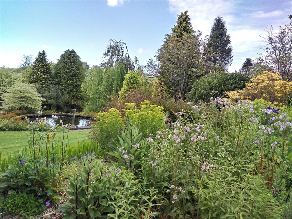 Lush spring perennials in the foreground with the formal round pond behind, showing a stretch of lawn between and mature trees beyond.
