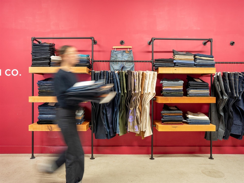 Person walking past shelves of neatly folded denim against a bold red wall at Hiut Denim. Various jeans styles and colours are displayed, with one pai