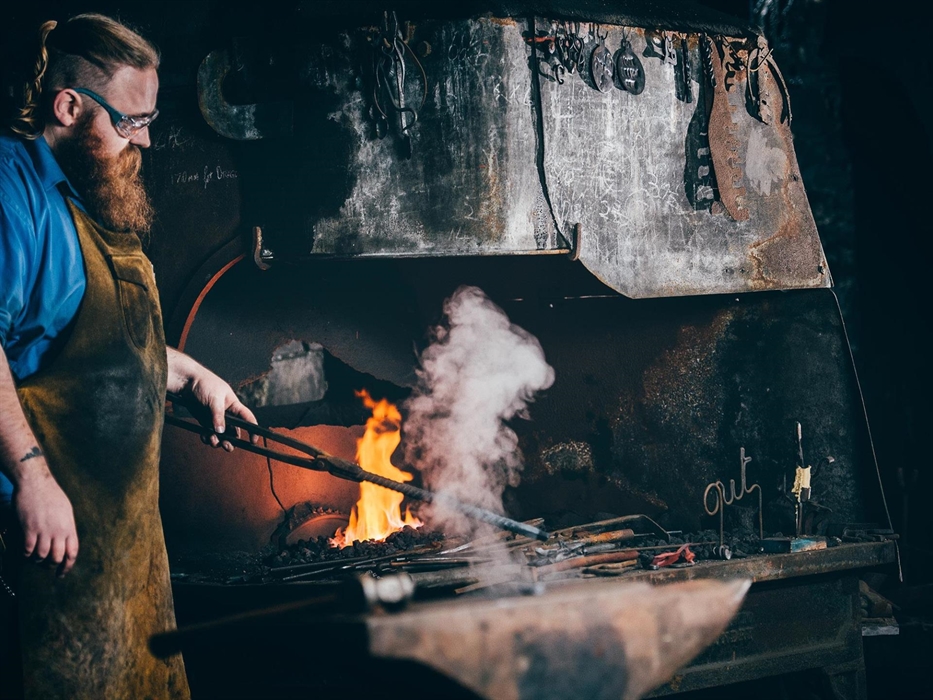 A blacksmith is dressed in a blue shirt and leather apron. He stands in front of a forge which is burning brightly. He holds a long metal rod that's j