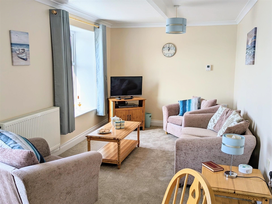 A photograph of the living room in apartment 5 showing the three piece suite, smart TV and beach coloured themed decor including duck egg blue curtain