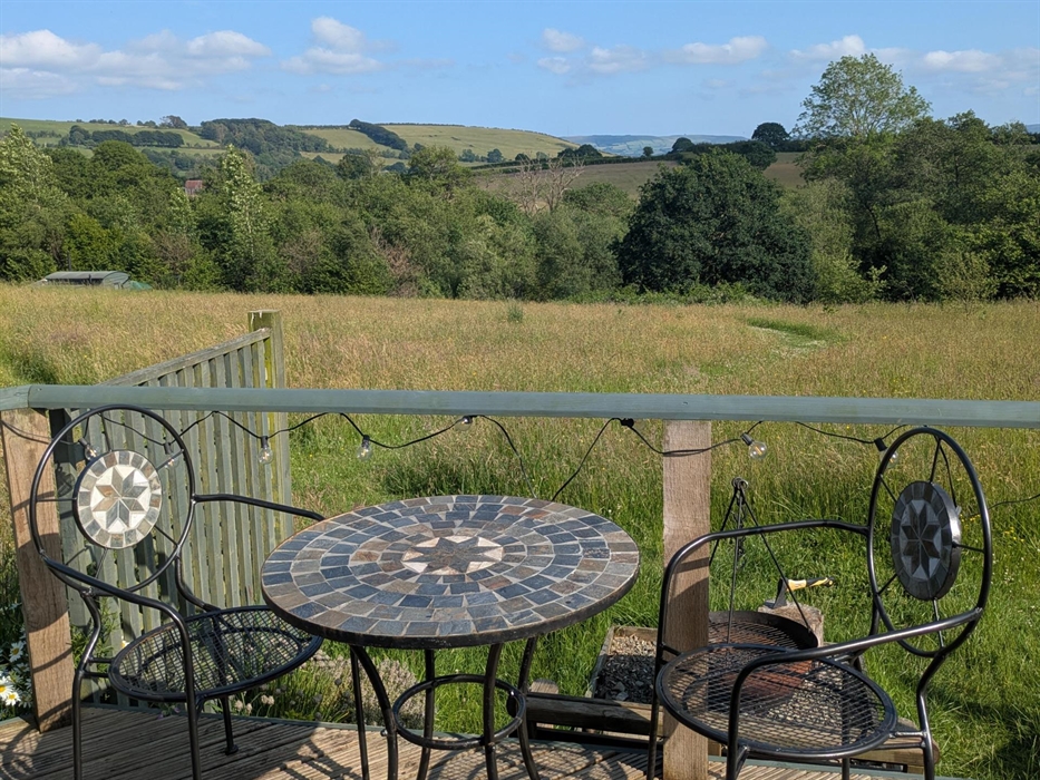Metal and stone mosaic outdoor table and chairs on the wood deck with a view over the wildflower meadow to the trees round the lake and the hills beyo