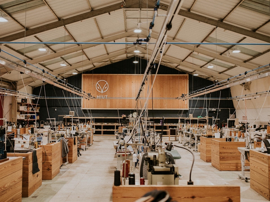 Interior of the Hiut Denim factory showing rows of wooden workstations with sewing machines under a high, well-lit ceiling. A large Hiut logo is displ