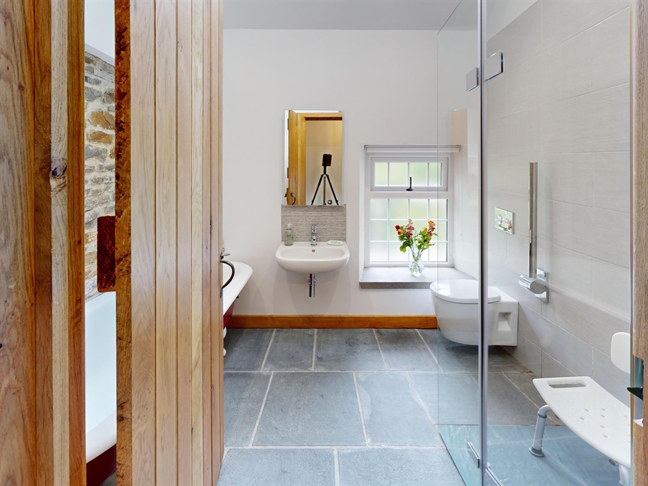 A bright bathroom with a walk-in shower, shower seat, wall-mounted toilet, sink, and a window with a vase of flowers, hand rails