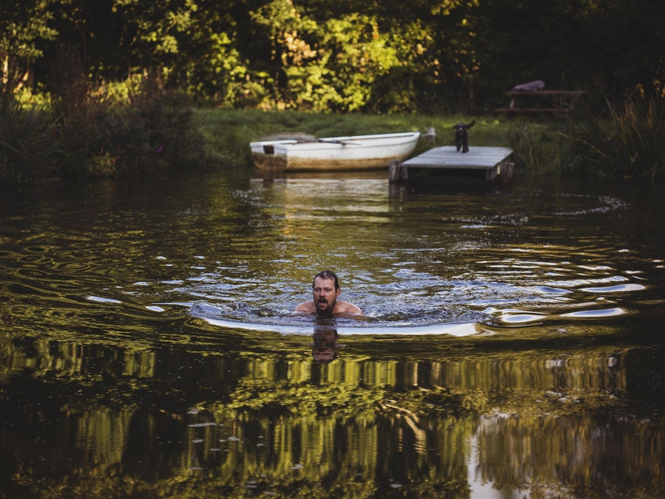 A man swims in the foreground.  the scene is of a pond with a jetty in the distance on which stands a dog with a rowing boat just seen.