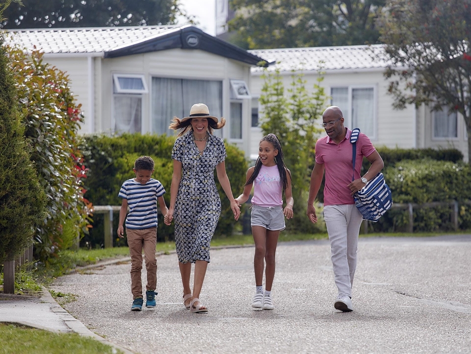 A family of 4 are walking alongside a row of caravans. The father is carrying a picnic bag.
