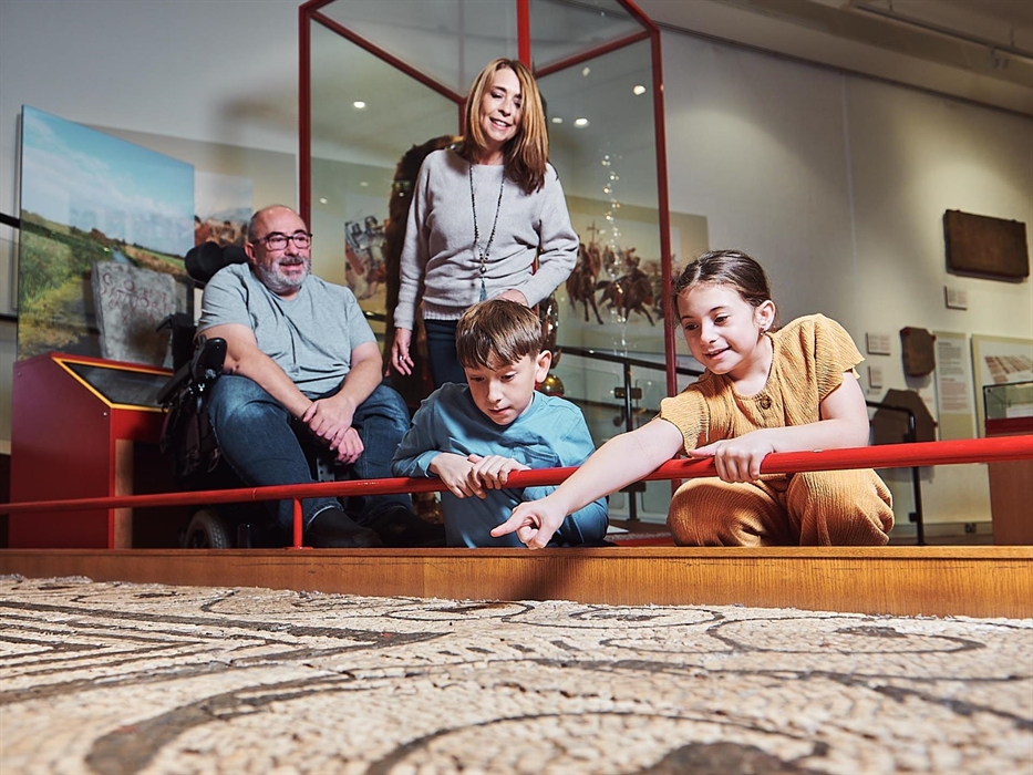 a young girl and boy, with mum and dad are in the museum gallery. the children are crouched down looking at a large Roman mosaic on display at floor l