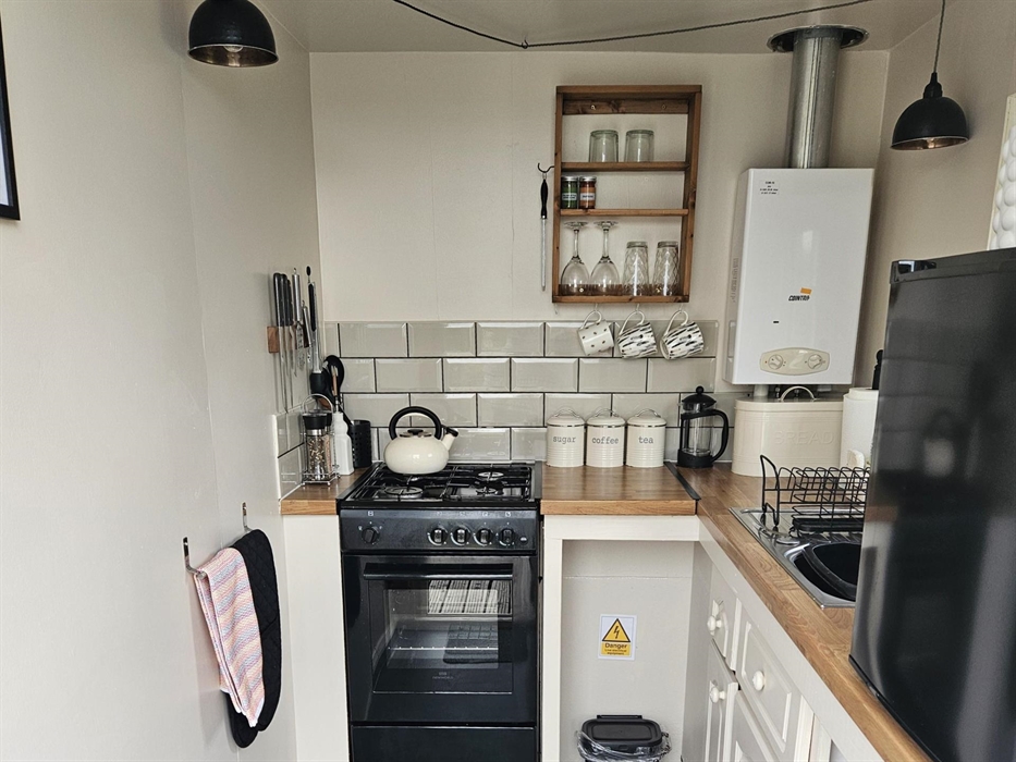 Black gas stove with hob top kettle, boiler, sink and black counter top fridge.Magnetic knife rack on wall. Utensils, tea, coffee,sugar canisters, cof