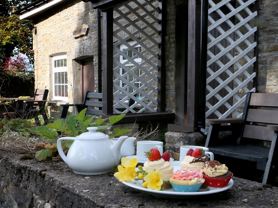 A plate of colourful cupcakes and a white teapot set on a stone wall outside a rustic stone cottage with outdoor seating and trellis.