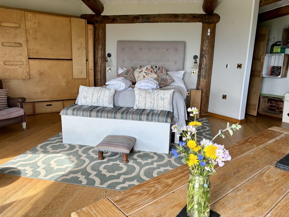 Inside Saffir Lodge, warm light fills a rustic open-plan space with wooden beams, soft furnishings and floral touches. A cozy armchair and natural tex