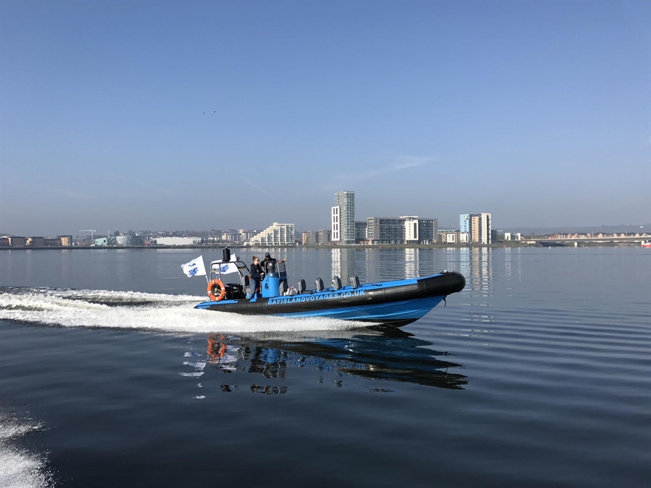 Blue Rib boat crossing Cardiff Bay with trailing wake behind it.  View of buildings in the background.