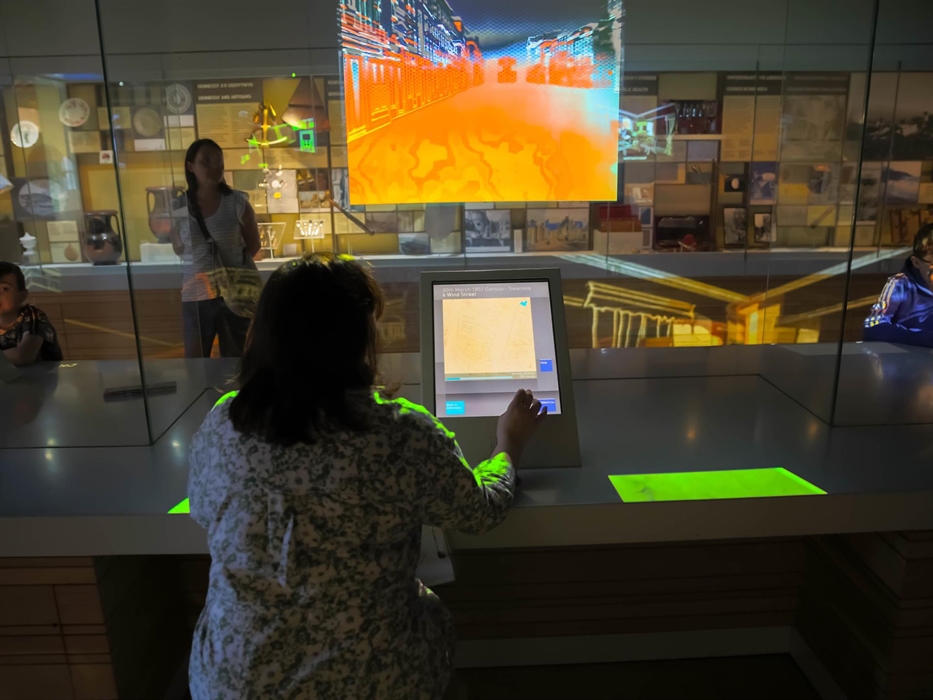 a lady sits at a large desk. on it is a computer screen that she touches. There is another large display screen above her on a glass wall. Through the
