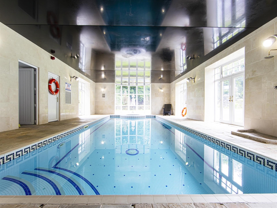 Bright and luxurious indoor pool at Plas Cilybebyll, featuring tiled walls, Grecian-style mosaic edging, and floor-to-ceiling windows overlooking the