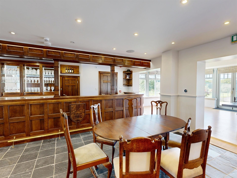 A warmly lit traditional bar with wood paneling, glassware on display, and elegant dining chairs at Plas Cilybebyll manor house.
