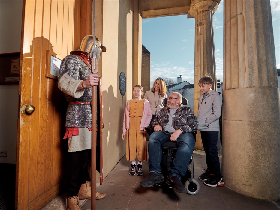 A man in gladiator costume stands to the left of the picture. He is meeting a family at the front entrance of the museum. It is a grand entrance with