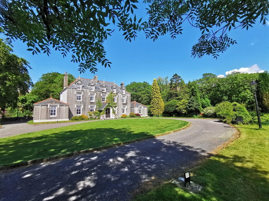 Striking exterior view of Plas Cilybebyll manor house, surrounded by mature trees, landscaped gardens, and a sweeping driveway under a clear blue sky.