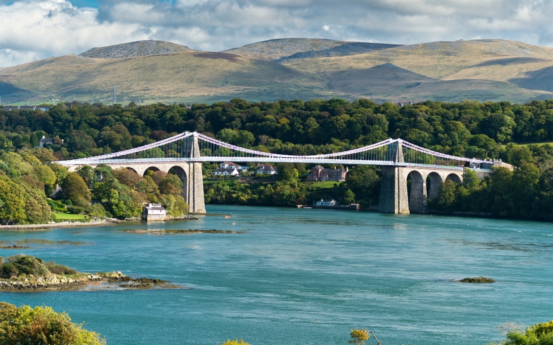 A suspension bridge with stone pillars spans a wide, blue river. Lush green trees and hills form the backdrop under a partly cloudy sky.