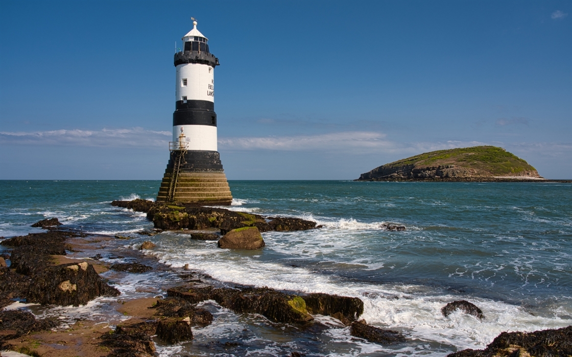 A black and white striped lighthouse stands on a rocky shore with waves crashing. In the distance, a small, green island is visible under a clear blue
