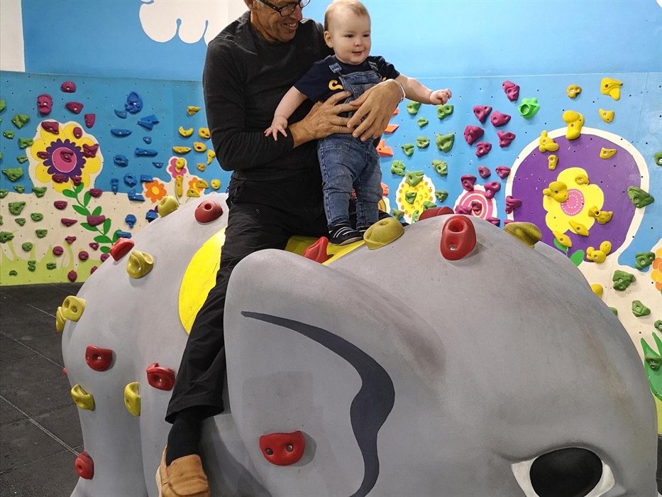Grandad and toddler grandson sit on a manufactured elephant with climbing holds on it, and a little colourful climbing wall behind them in the play ar