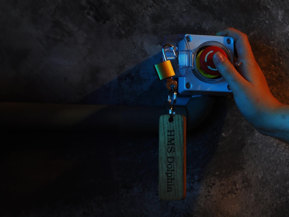 A hand is poised over an emergency stop button with a lock and key hanging off with a piece of wood inscribed HMS Dolphin