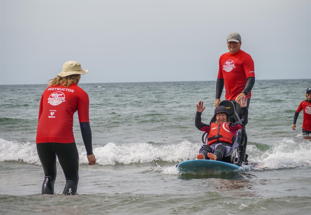 A disabled lady on a seated surfboard on a wave in the sea with her arms in the arm and smiling, with a surf instructor surfing on the board with her